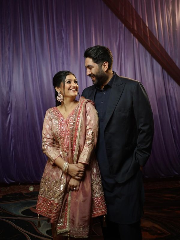 The couple smiling at each other in front of a purple curtain.